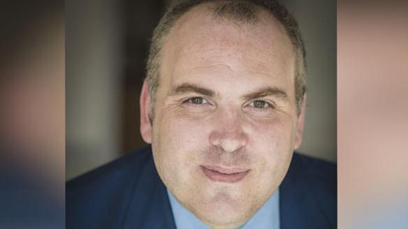 Man looking directly at the camera. He is wearing a yellow tie with a light blue shirt and a dark blue suit.