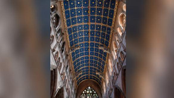 The vaulted ceiling at Carlisle Cathedral. It is divided into squares surrounded by a golden frame, each square features a starry, blue sky.