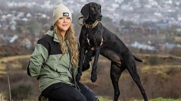 Sophie is kneeling beside Gwen and holding her up so that her two front paws are off the ground. Sophie is wearing a winter hat and coat and has long curly blonde hair. Gwen is all black with a white patch on her chest. She was long floppy ears and her shortened tail is visible. The pair are on a grassy hilltop overlooking a town 