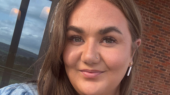 A selfie picture of a woman with long brown hair, wearing a blue top, and silver earrings, nose ring and heart-shaped necklace. She is smiling and looking at the camera, and is stood in front of the wall of a house which has red brick and large french-door style windows. It is a head shot of her.