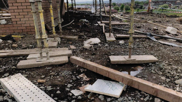 The ground covered in building materials in a unorderly state. The bottoms of scaffolding poles are on the group and a partly constructed house stands on the left.