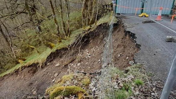 A close up of the landslip. A section of the carriageway has crumbled away leaving a steep slop of brown earth running to the left of the screen. A metal fence has been erected around the area to prevent people from coming too close to the slope.