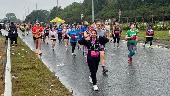 Miss Ahmed among dozens of runners taking part in Great North Run on a rainy day. She is wearing black running gear and is smiling and giving a thumbs up to the camera. 