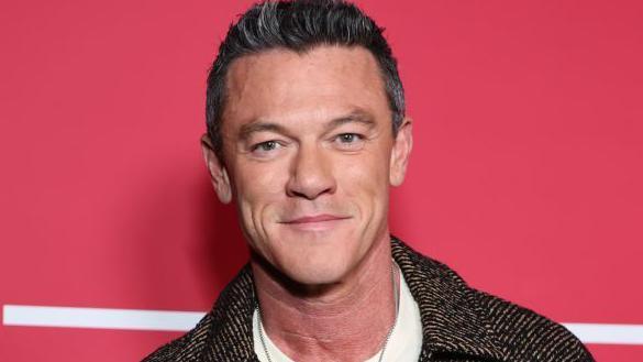 Luke Evans smiling at the camera. He is standing in front of a red background with a white line just behind him. He is wearing a dark brown tweed jacket and a silver necklace. 