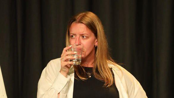 Ruby Tapp wearing a black top with a white shirt over the top of it. She is drinking from a glass which is in her left hand. She has shoulder-length brown hair and is looking to the left.