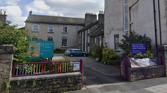 The Quaker Tapestry Museum in Kendal. It is an old Georgian house with two signs in front advertising the museum and a sign reading 'welcome to Quaker meeting house'. The entrance to the car park has coloured railings.