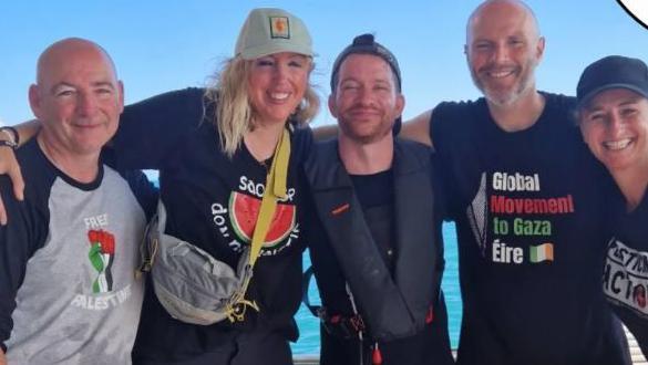 Three men and two women post for a photo on board a ship on a sunny day.  They are all smiling and have their arms around each other's shoulders.  All of them are wearing different t-shirts with pro-Palestinian logos. 