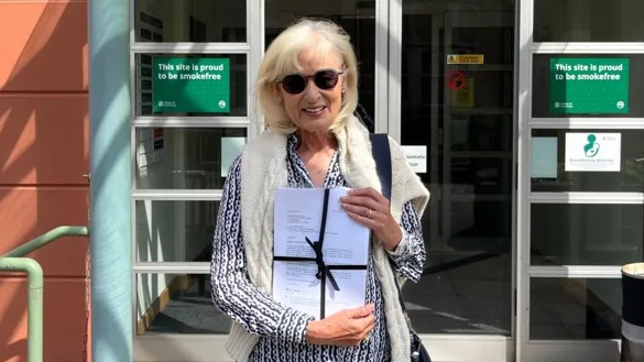 Andrea Lewis outside a government building called Sir Charles Frossard House, holding a petition wrapped in a black ribbon. She has white hair and is wearing a pattered shirt and sunglasses, and has a white jumper over her shoulders.