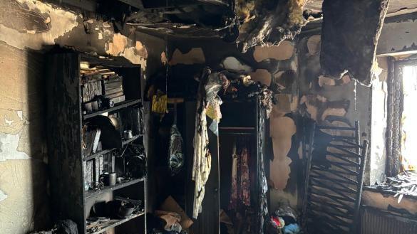 A bedroom destroyed by a fire. Shelves and wardrobes are blackened by fire damage, including their contents. The ceiling has partially collapsed.
