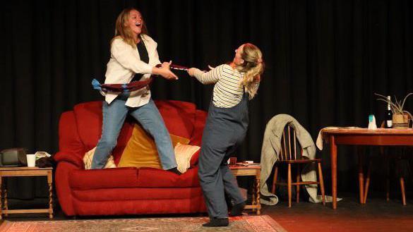 Two women on stage pulling a scarf between them. One woman is stood on a red sofa, and the other woman is stood on the floor. There is a dining table on the right with a chair pulled out. To the right of the sofa, there is a wooden side table with a white cup on it.