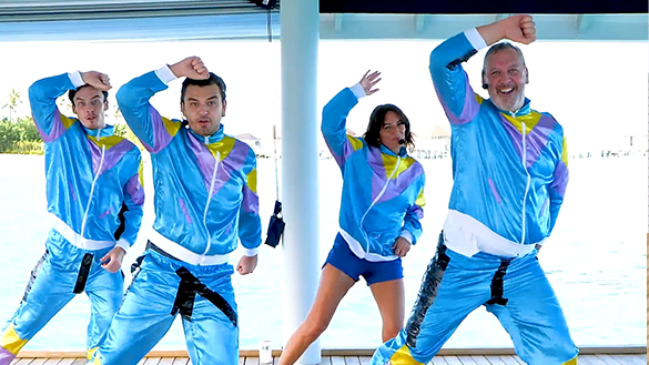 Travis, Harrison, Davina McCall and Paul, all dancing, with one arm up, in tracksuits. Davina has a headset on.