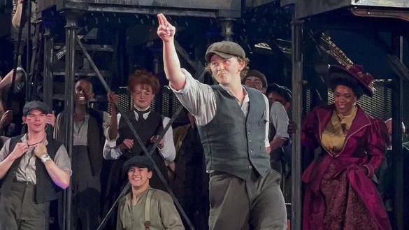 A man wearing a flat cap, waistcoat and grey shirt pointing up towards the ceiling, with other actors in the background wearing similar attire.