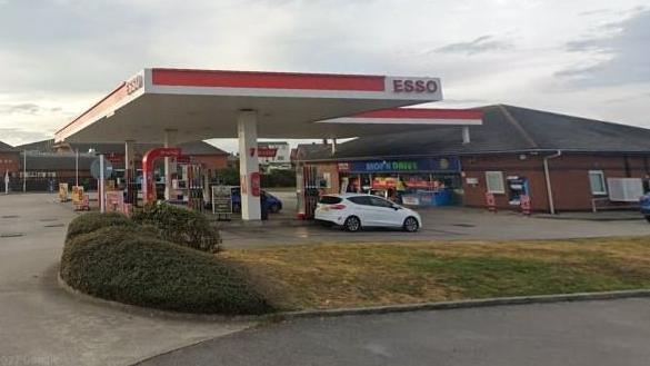 An Esso petrol station with several cars filling up. There is also a convenience store in the same building where drivers are paying for fuel. 