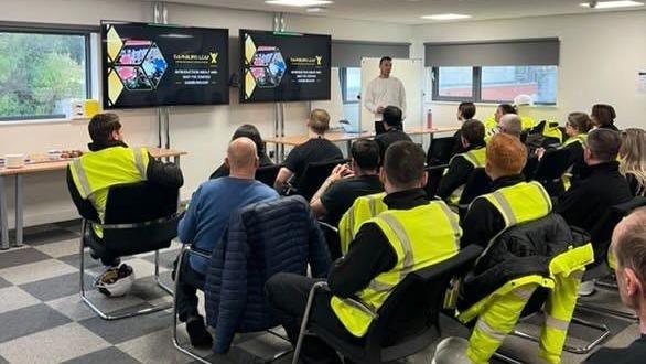 Colin in a white top at the front of a classroom of workers looking at slides. Many of them are wearing hi vis tops.