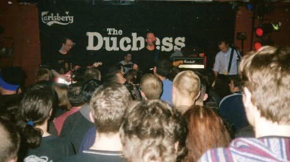 An old photograph showing a crowd of people watching a band play on a stage with a black sign behind with white writing which says The Duchess.