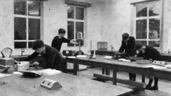 A black and white shot of a science classroom in a school. Four boys work at two long benches, which have power outlets and other pieces of equipment.