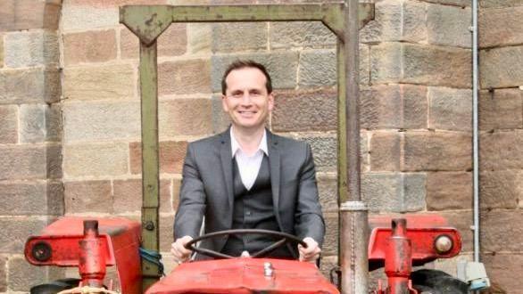 Tim Connors wears a dark suit and white shirt and is sat on a red tractor holding the wheel in his hands