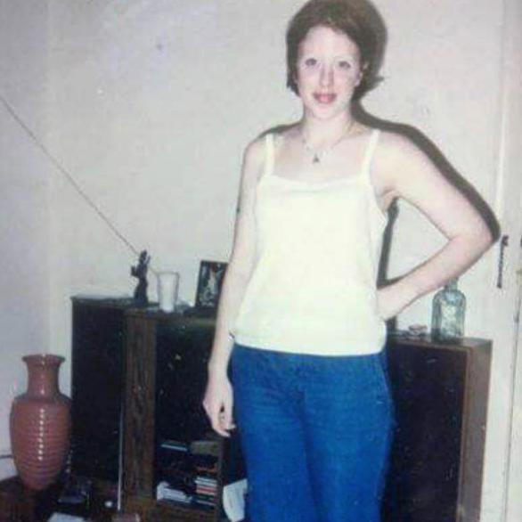 The Blessed Madonna as a 20-year-old, she has short brown hair and is wearing a yellow vest and blue jeans and is stood in front of a home stereo system.