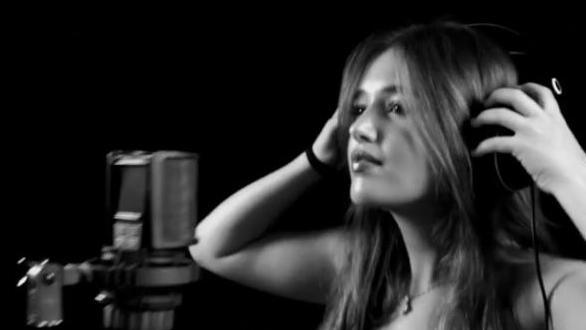 A black and white photo of a woman with dark hair down past her shoulders is standing in front of a studio microphone and holding headphones on her head