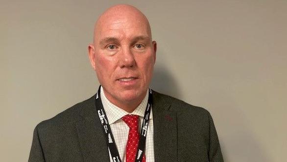 Phil Jame is wearing a grey suit jacket with cream checked shirt and a red tie with white spots on it and had a black lanyard around his neck.