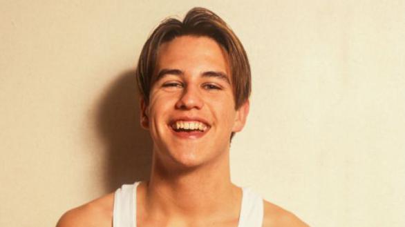 Anthony Kavanagh as a young star grins for a photo shoot. He has light coloured hair and is wearing a white vest top.