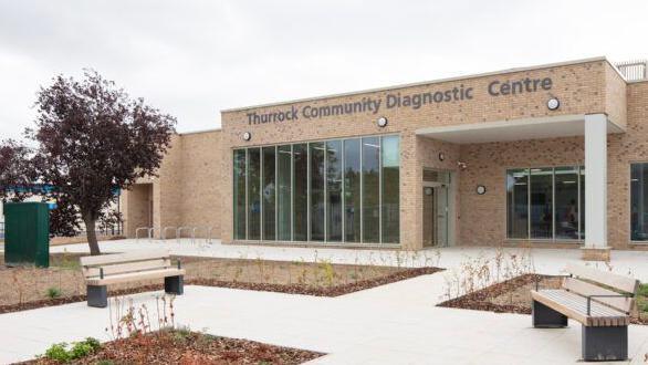 The community diagnostic centre in Thurrock. It is made of cream bricks and has a large window at the front. White paving leads to the entrance and there are wooden benches. There are also flower beds with small saplings growing from them.