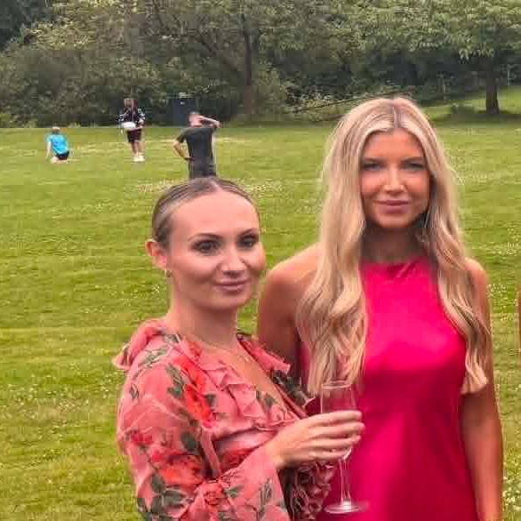 A woman with brown hair tied up is wearing a pink, floral dress and holding a glass of bubbles.  She is next to a woman with long blonde hair weaeing a bright pink dress.  They are outside, stood on some grass.