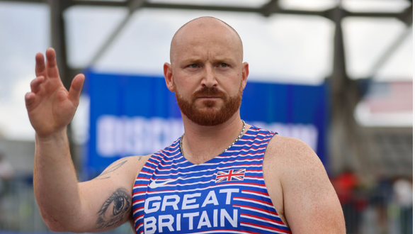 A man with a bald head and red beard wearing a blue sports vest holds his right hand up. 
