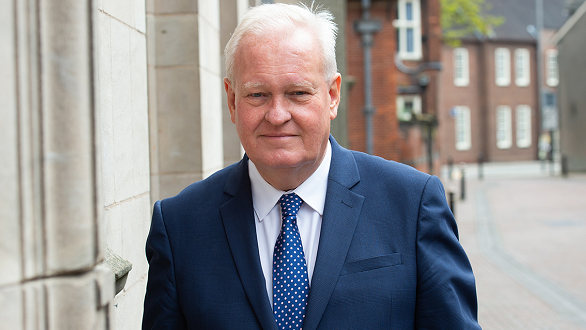 Councillor Ian Parry is wearing a white shirt and blue suit, with a blue tie that has white spots on it. He is stood in a street, with red brick buildings in the background.