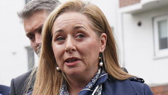 BBC Spotlight NI reporter Jennifer O'Leary outside the High Court in Dublin. She has long, straight auburn hair and is wearing a navy trench coat over a white blouse. She is also wearing a multi-coloured blue and white scarf.