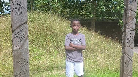 Othniel Adoma standing in front of a grass mound between two wooden pillars.