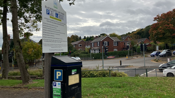 A pay and display machine in a car park. A main road is visible behind it, with a residential estate on the other side.