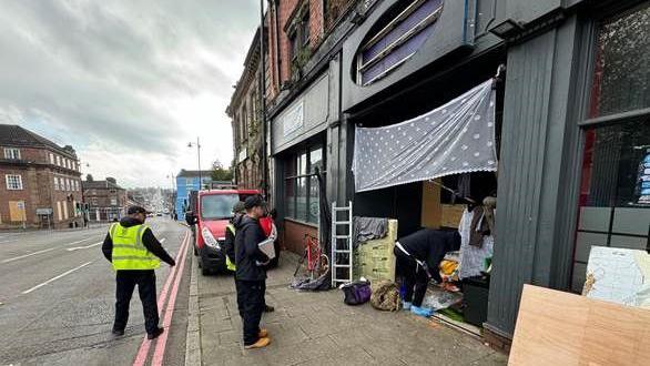 Council workers remove a homeless camp from a doorway of a vacant restaurant - three people are standing watching another person clean up items from the doorway, which has a towel stretched across the top. 