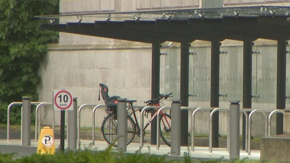 A metal and glass bike shelter with metals railings to prop bicycles against. One bike is in the shelter. Behind the shelter a grey building