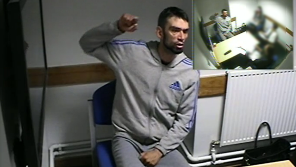 A man with dark hair and a beard wearing a grey track suit raised his right hand while sitting in a police interview room.