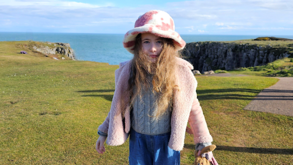 Gwen Harries looking at the camera. She is stood on grass at the edge of a cliff with the sea behind her. She is wearing a pink coat and a pink and white spotty hat.