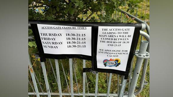 Two paper signs taped to gate. One reads: "Access Gate Closure Times 18:30 - 21:15', while the other reads: 'The access gate leading to the main area will be closed between the hours of 18:30 and 23:15'