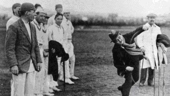 A black and white photo of a man wearing a coat, scarf and hat bowling a cricket bowl. A group of men are stood to the left if the photo in cricket whites.