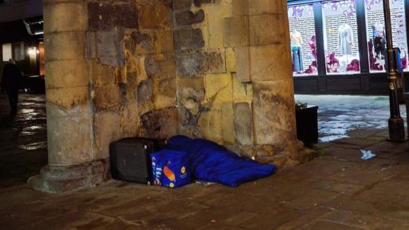 A person in a blue sleeping bag in Shrewsbury. They are against an old wall and there is a shop lit up in the background. 