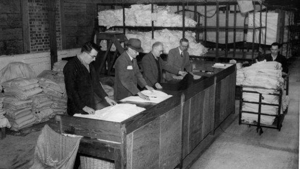 Four men are lined up behind a wooden desk. Each is folding a piece of uniform. A man is wheeling a trolley piled high with white shirts past the desk. In the background are large wooden shelves filled with folded white garments and there are piles of the same on the floor behind the men.