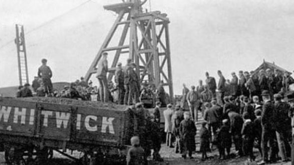 A black and white photo, showing a coal wagon with the word Whitwick, on the side in large letters. It stands in front of a pit head structure.  A crown of men stand around it