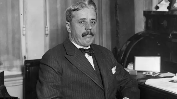 A black and white picture from the early 20th Century of a man sitting on a chair in an office. He is wearing a pin-striped suit and a bow tie. He is looking into the camera but is not smiling. 