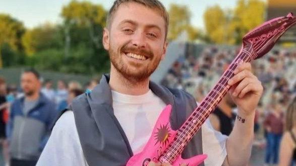 Kian Broadhead is pictured at what appears to be a music festival. He is holding a small pink inflatable guitar and is smiling. Kian is wearing a white T-shirt and he has a grey sweatshirt draped over his shoulders. He has short reddish-blonde hair and a short beard.
