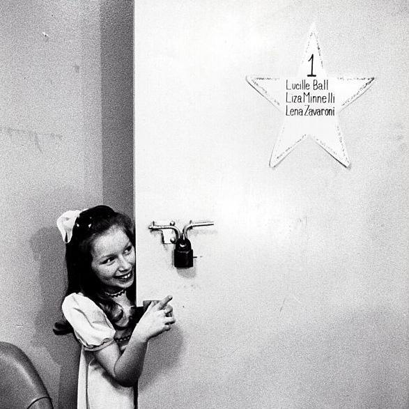 A black and white photo of Lena, in her trademark pinafore dress and bow, peeking out from behind her dressing room door. A star on the door lists its occupants - Lucille Ball, Liza Minelli and Lena Zavaroni.