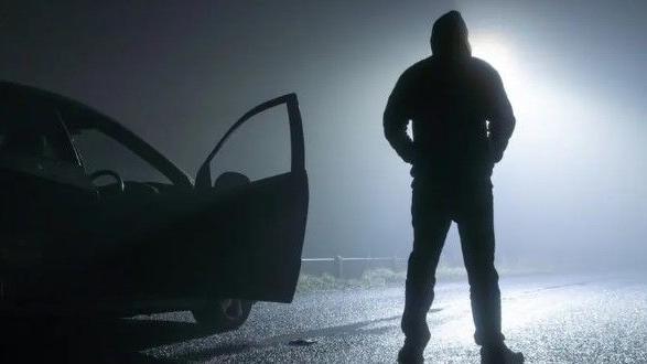 A man in silhouette stands in the middle of the road beside a car. 
The car door is open and it looks like he is waiting for someone. 