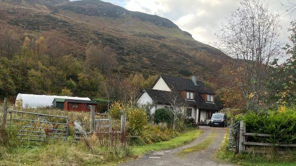 A house is at the end of a track. There is a van parked outside. Next to the house are sheds and a plastic horticulture tunnel, and a hill in the background.