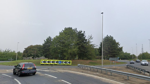 A roundabout with a neon arrow sign in its centre. There is one car approaching the roundabout and three cars coming off the roundabout and driving on the opposite carriageway. 