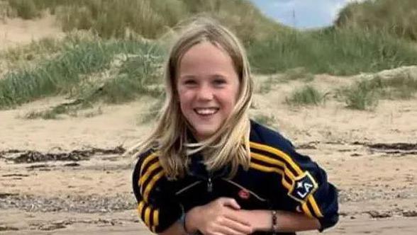 Evan Rothwell, aged 11, with long blond hair sits on the shoulders of a statue on a beach and smiles at the camera.