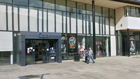 The exterior of Durham's Gala theatre, a glass fronted building with black steel posts and signage spelling out Gala against a grey entrance. Posters for upcoming shows are visible. A man and a woman are walking in front of the building.