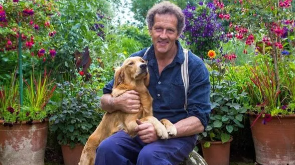 Monty Don, a middle-aged man with short dark curly hair, sits in a garden surrounded by large pots filled with plants and flowers. He is holding a golden retriever dog, which has its front paws on his lap.